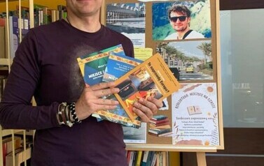 Michał Szulim stoi trzymając trzy swoje książki. Za Nim jest tablica korkowa z grafiką o Tygodniu Bibliotek.