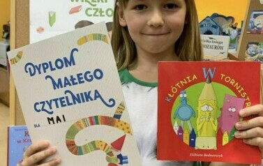 Dziewczynka w ciemnych blond włosach pokazuje sw&oacute;j nowy dyplom i wypożyczoną książeczkę. Za nią stoi korkowa tablica z plakatem promującym akcję czytelniczą.