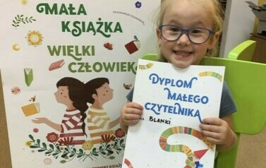 Uśmiechnięta dziewczynka w okularach i blond włosach pokazuje sw&oacute;j nowy dyplom. Z lewej strony stoi tablica z plakatem promującym akcję czytelniczą.