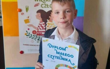 Chłopiec w blond włosach pokazuje sw&oacute;j dyplom. Za nim stoi metalowa tablica z plakatem dotyczącym akcji "Mała Książka Wileki Człowiek"