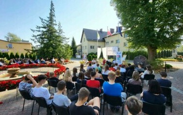 Widok z daleka na siedzącą publiczność i czytających Radnych. Po środku jest baner, po lewej stronie firanki,a po prawej rolka z logiem biblioteki. Po środku jest fontanna otoczona kwiatami.