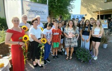 Przy banerze stoi grupa uczni&oacute;w z kierowniczką biblioteki i Radnymi, kt&oacute;rzy trzymają słoneczniki.