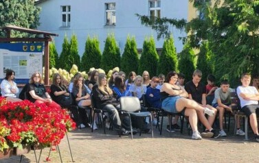 Skiedząca publiczność młodzieżową,a obok kwiaty czerwone przy fontannie.