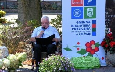 Radny czyta tekst "Kordiana" wśr&oacute;d kwiat&oacute;w,a po prawej stronie stoi rolka z logiem biblioteki.