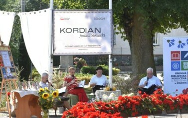 Czterech Radnych siedzi przy stoliku czytając "Kordiana". Widać baner, firanki, kwiaty czerwone przy fontannie, słoneczniki i rolkę z logo biblioteki.