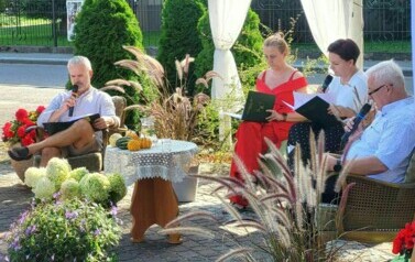 Czterech Radnych siedzi przy stoliku wśr&oacute;d roślin i firanek dekoracyjnych czytając "Kordiana". 