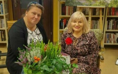 Pisarka przy stoliku podpisuje czytelniczce książkę. Na stoliku przed nimi stoją dwa kwiaty doniczkowe i leżą małe dynie. Za nimi stoi duży ekran z wyświetlonym plakatam o spotkaniu.