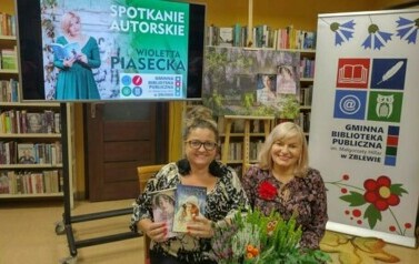 Kierowniczka biblioteki siedzi razem z pisarką. Trzyma dwie książki autorki. Uśmiechają się. Przed nimi na stoliku stoi duży kwiat doniczkowy.