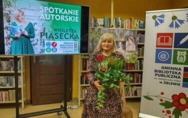 Na środku stoi pisarka z kwiatami, z prawej strony jest rolka z logiem biblioteki,a z lewej duży ekran z wyświetlonym plakatem o spotkaniu.