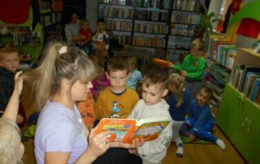 Dzieci siedzą na dywanie i słuchają bajki czytanej przez bibliotekarkę.