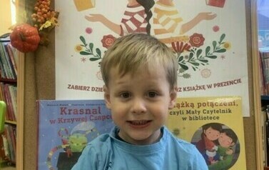 Chłopiec w blond włosach i w niebieskiej bluzce stoi trzymając nową książkę  z wyprawki i kartę na naklejki. Za nim stoi korkowa tablica z plakatem promującym akcję.