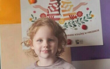 Dziewczynka w blond i lekko kręconych włosach stoi trzymając swoją nową książkę z wyprawki czytelniczej. Za nią stoi metalowa tablica z plakatem promującym akcję.