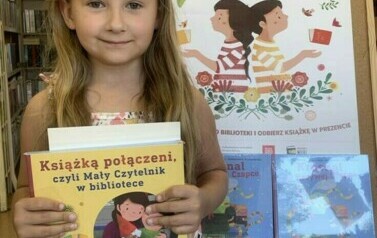 Dziewczynka w długich blond włosach stoi trzymając nową książkę z wyprawki. Za nią stoi korkowa tablica z plakatem i książkami promującymi akcję.