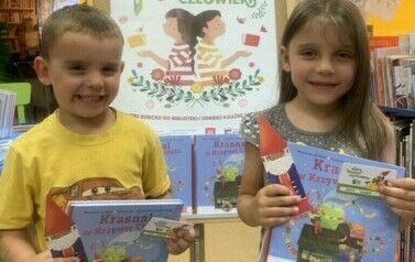Dziewczynka z chłopcem stoją pokazując swoją wyprawkę czytelniczą. Za nimi stoi tablica z plakatem dotyczącym akcji.