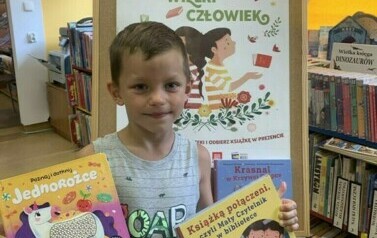 Chłopiec  w ciemnych włosach stoi trzymając nową książeczkę z wyprawki i z książeczką, kt&oacute;rą wypożyczył. Za nim stoi tablica korkowa z plakatem promującym akcję.
