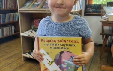 W bibliotece w Borzechowie stoi dziewczynka w blond włosach z nową książeczką i zakładką do niej.