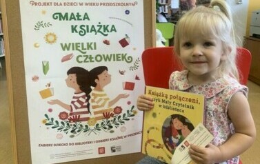 Dzieczynka w blond włosach i w kitku siedzi na krzesełku i trzyma swoją nową książeczkę i zakładkę do niej. Za Nią stoi tablica korkowa z plakatem promującym akcję Mała Książka Wielki Człowiek