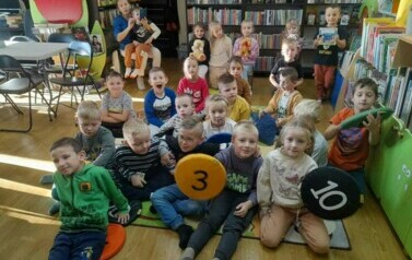 Dzieci siedzą na dywanie i słuchają bajki, kt&oacute;rą czyta im bibliotekarka. Dw&oacute;ch chłopc&oacute;w trzyma w rękach siedziska.