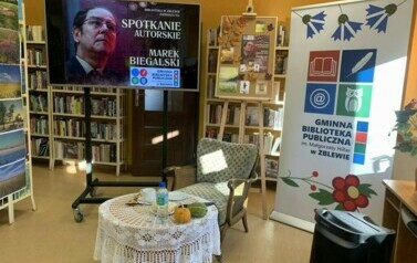 Po środku stoi okrągły stolik z fotelem. Po prawej stronie stoi rolap z logiem biblioteki, a po lewej stronie stoi duży ekran z wyświetlanym plakatem ze spotkania.