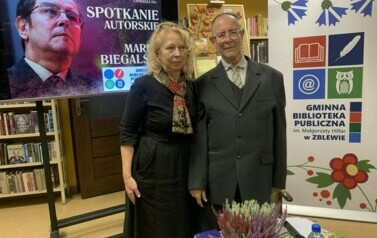 Autor wierszy stoi z czytelniczką przy stoliku. Po lewej stronie stoi duży kran z wyświetlonym plakatem o spotkaniu, a po prawej stronie stoi rolo z logiem biblioteki.