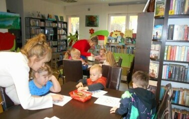 Przy stołach dzieci wykonują prace plastyczne, a ich Pani i Pani z biblioteki są pochylone nad stołami i im pomagają.
