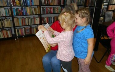 Dwie dziewczynki stoją przy Pani bibliotekarce i wyklejają jakieś elementy w książeczce.