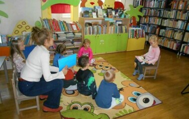 Dzieci wraz ze swoją Panią siedzą na dywanie,a z przodu siedzi na krzesłku dziewczynka i patrzy na jakąś kartkę, kt&oacute;rą trzyma w ręku.