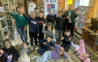 Zdjęcie grupowe dzieci w pozycji stojącej i leżącej na dywanie wraz ze swoją Panią i Panią z biblioteki. Machają rękami uśmiechając się.