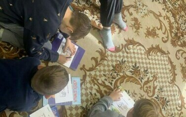 Dzieci w pozycji siedzącej i leżącej rozwiązują na dywanie krzyż&oacute;wki.