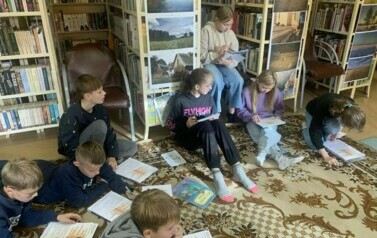 Dzieci siedzą na dywanie i rozwiązują na papierze krzyż&oacute;wki.