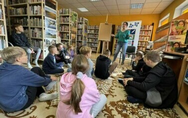 Dzieci siedzą na dywanie, a z przodu stoi bibliotekarka i ma rozłożone ręce w geście tłumaczenia zasad funkcjonowania biblioteki.