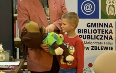Pan w r&oacute;żowej marynarce i czerwonym kapeluszu trzyma leżącego misia pod brodą, a chłopiec stoi przy tym i się patrzy.
