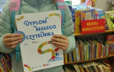 Dziewczynka w upiętych blond włosach pokazuje dyplom. Za nią stoją regały z książkami w bibliotece w Borzechowie.