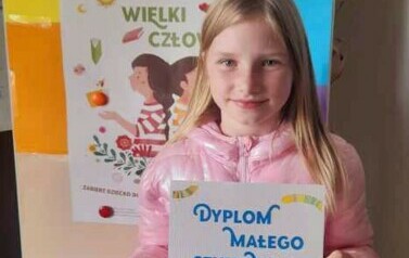 Dziewczynka w blond wlosach i w r&oacute;zowej kurtce pokazuje sw&oacute;j dyplom. Za Nią stoi metalowa tablica z plakatem dotyczącym akcji.