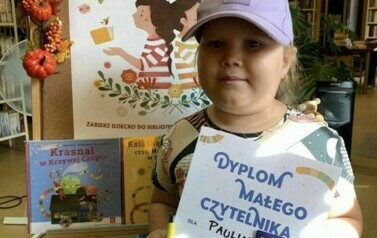 Dziewczynka w fioletowej czapce z daszkiem pokazuje sw&oacute;j dyplom i kartę Małego Czytelnika. Za nią stoi korkowa tablica z plakatem i książkami dotyczącymi akcji.
