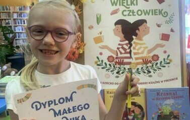 Dziewczynka w upiętych blond włosach pokazuje dyplom i długopis. Uśmiecha się. Obok stoi korkowa tablica z plakatem i książkami dotyczącymi akcji.