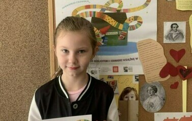 Dziewczynka w blond włosach i ciemnej bluzie pokazuje sw&oacute;j dyplom, za nią jest korkowa tablica z plakatem dotyczącym akcji i innymi ciekawostkami.