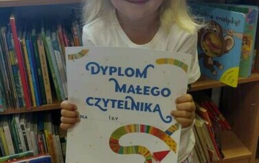 Dziewczynka w blond włosach z r&oacute;źową opaską pokazuje sw&oacute;j dyplom, za nią stoi regał z książkami w bibliotece w Borzechowie.