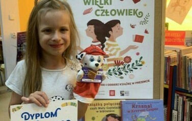 Dziewczynka w dłuższych blond włosach trzyma w rękach dyplom i kukiełkę, obk stoi korkowa tablica z plakatem i książkami dotyczącymi akcji.