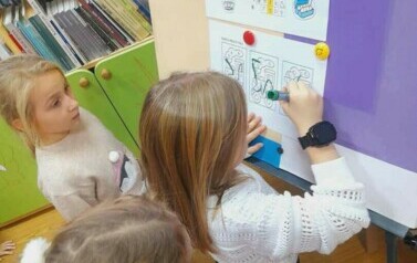 Dziewczynka w blond włosach przy tablicy  łączy lierki w wydrukowanych wagonach aby powstalo słowo "Przepraszam".Obok niej są inne dzieci.