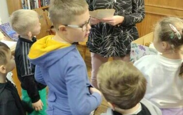 Dzieci stoją przy Pani pedagog, kt&oacute;ra trzyma w rękach stare kartki od książki.