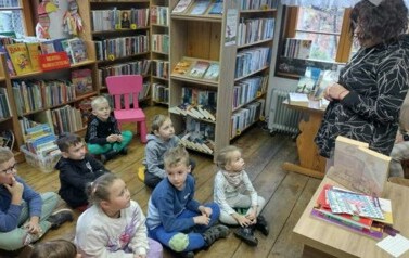 Dzieci siedzą na siedziskach w bibliotece i patrzą na Panią pedagog, kt&oacute;ra stoi z przodu i do nich m&oacute;wi.