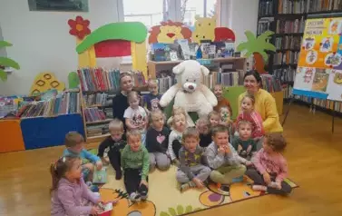 Dzieci siedzą na dywanie, po środku na krześle siedzi duży biały miś, a po bokach siedzą bibliotekarka i nauczycielka przedszkola.