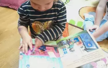 Dziewczynka siedzi na poduszce i ogląda papierową książeczkę.