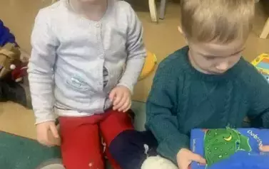 Chłopiec w blond włosach bawi sie pluszową książeczką obok niego siedzi dziewczynka.