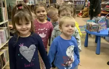 Uśmiechnięci dziewczynka z chłopcem stoją w parze trzymając się za ręce. Za nimi stoją inne dzieci w parach. Po prawej stronie stoi ich Pani.
