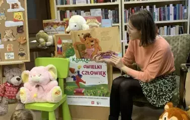 Pani siedzi na krześle i czyta bajkę, obok na krzesełku i stole leżą misie, na środku jest korkowa tablica z plakatem promującym akcję Mała Książka Wielki Człowiek.