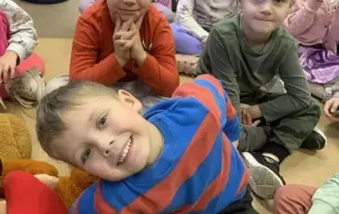 Dzieci siedzą na siedziskach, z przodu chłopiec siedzi pochylony w lewą stronę i się uśmiecha.