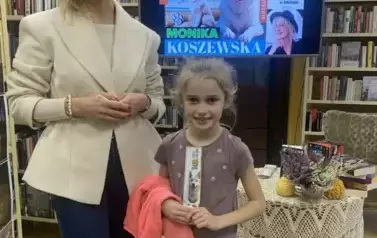 Dziewczynka w blond włosach stoi przy pisarce, ma na ręce przewieszoną bluzę i trzyma w rękach zakładkę do książki. Obie się uśmiechają.