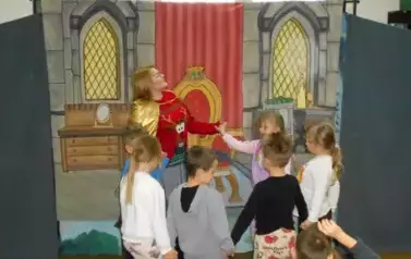Na tle wnętrza pałacu aktorka w przebraniu renifera ze złotą peleryną tańczy w k&oacute;łeczko z dziećmi z publiczności.